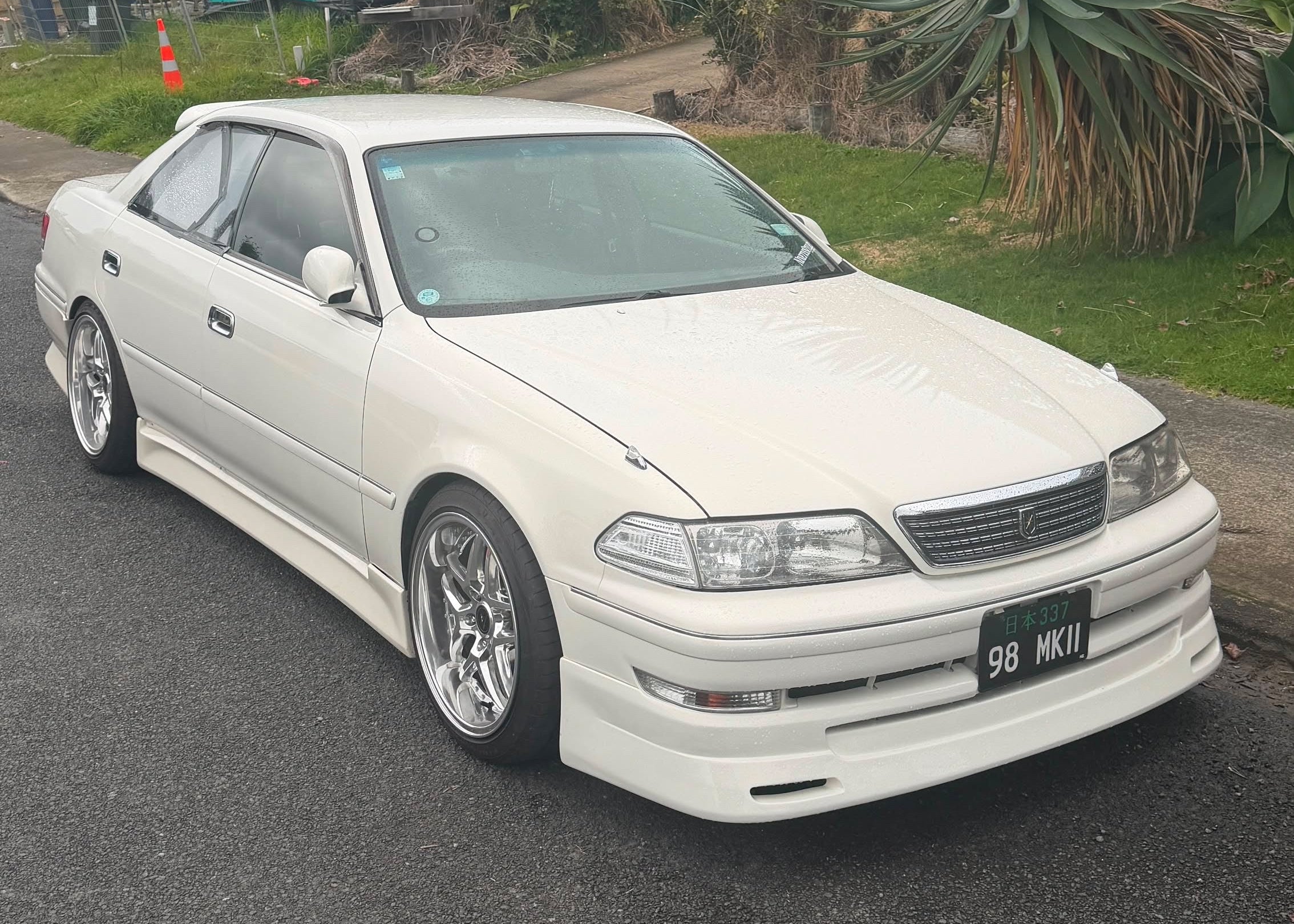 A white customized Toyota mark ii jzx100 with aftermarket chrome wheels, a lowered suspension, and a custom front bumper, parked on an asphalt road with grass and trees in the background.